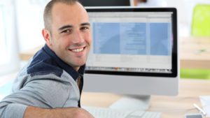 A male professional smiling looking into the camera while working on his office desktop